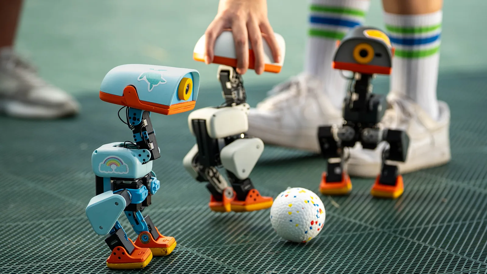 Three Microduck robots in sky blue, white and graphite on a sports court next to a ball, one of them being set down by hand